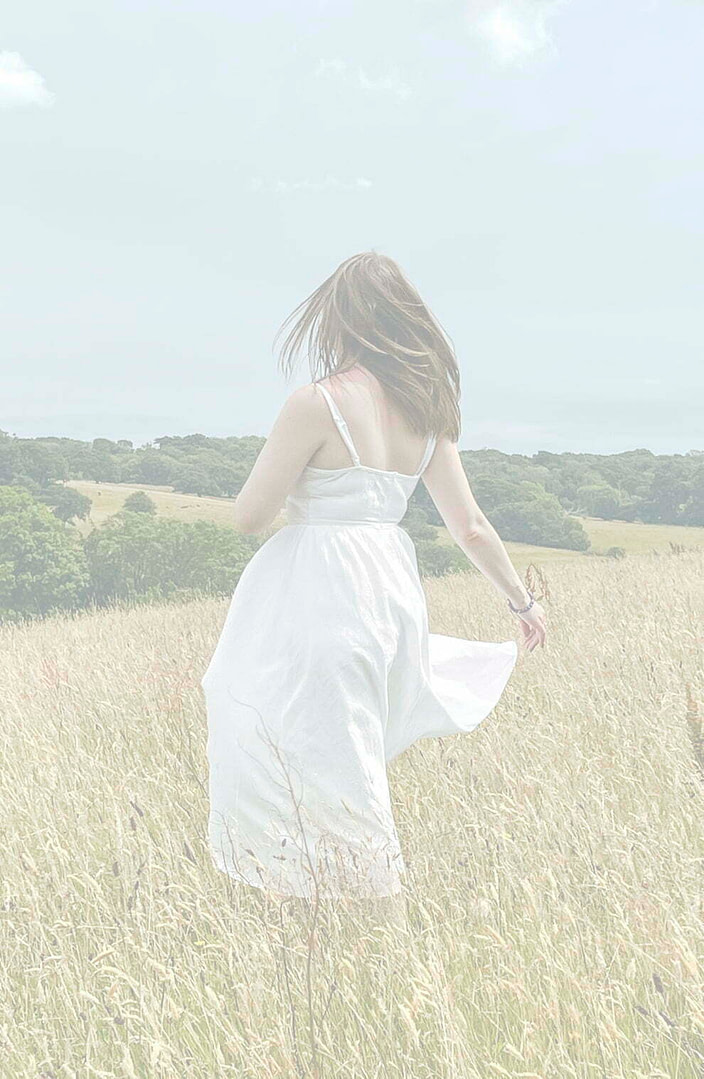 Female Empowerment Coach Louise Anne in a field of long grass in a white dress facing away from camera