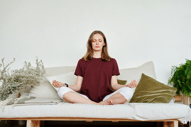 Woman sitting on sofa, eyes closed, in a meditation pose.